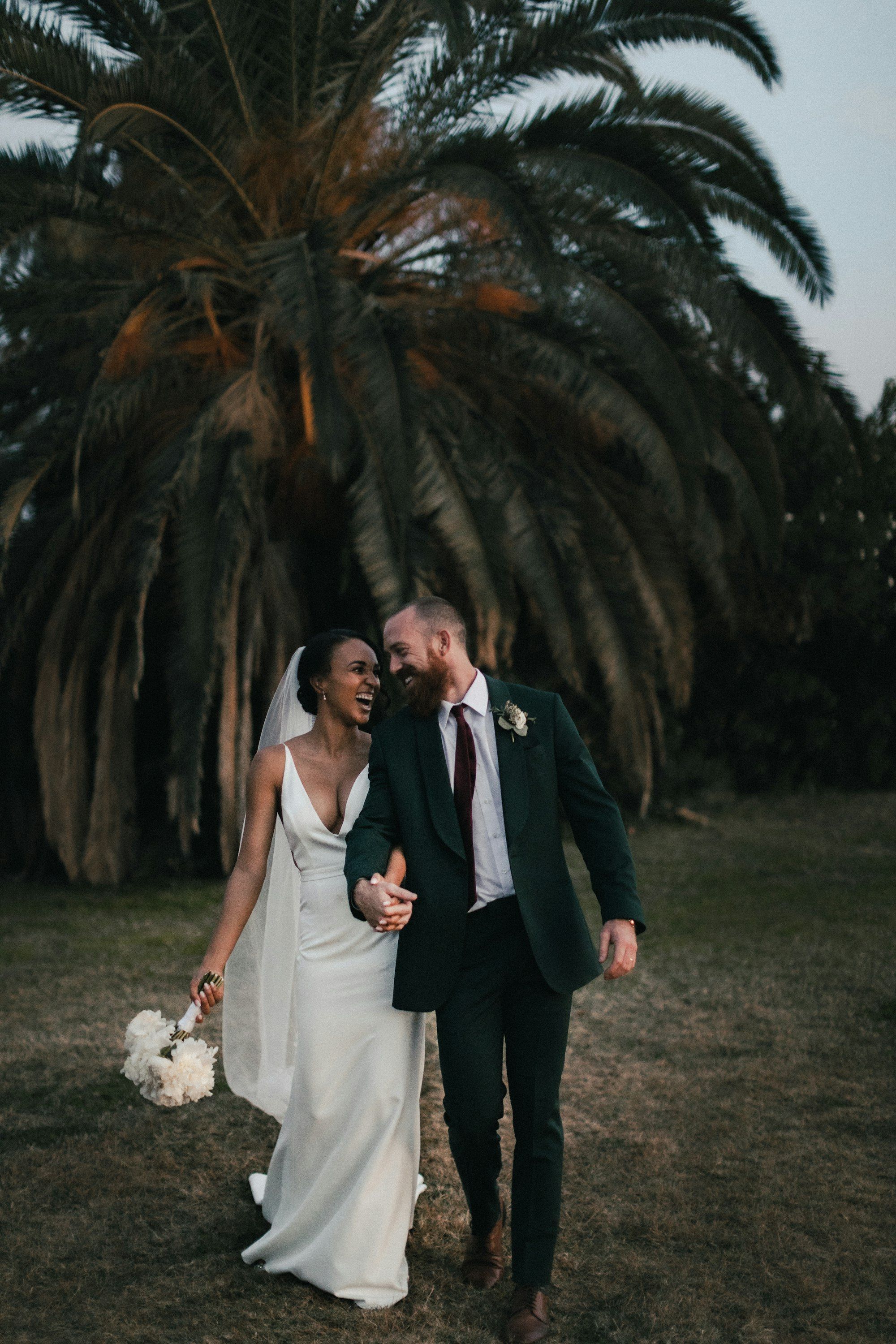 The newlyweds walking hand in hand beneath a palm tree at dusk